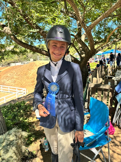 Lily Kagchelland, a young girl with a horse riding helmet and formal riding attire stands in front of a tree, holding a blue ribbon awarded for her performance at an equestrian event.